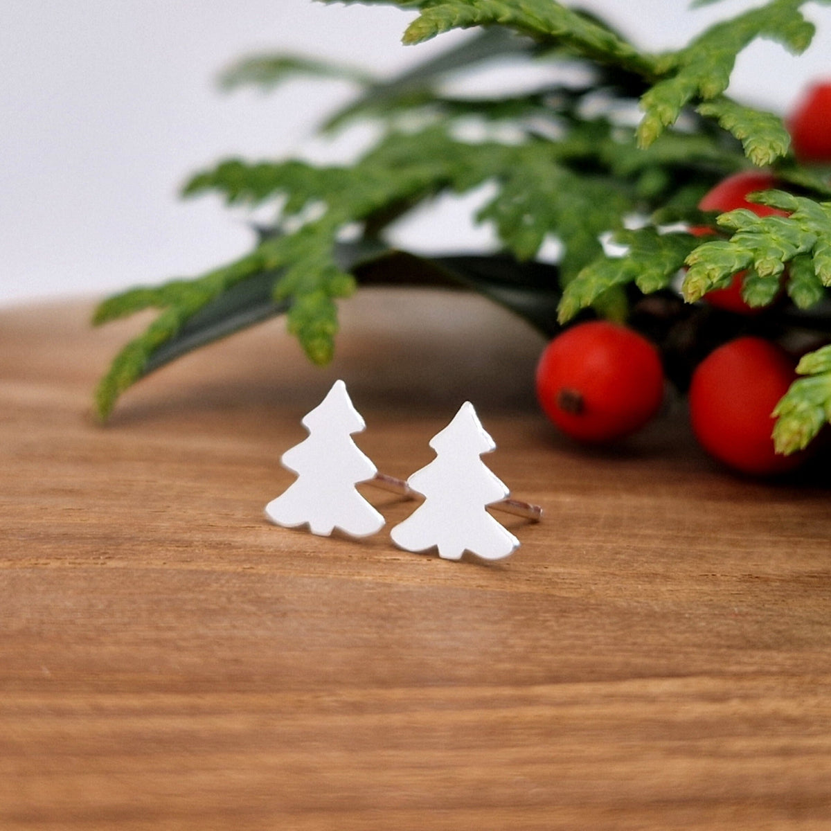 WeihnachtlicheTannenbaum-Ohrstecker aus 925 Silber in Nahaufnahme auf Holzteller mit Pflanzen mit roten Beeren im Hintergrund