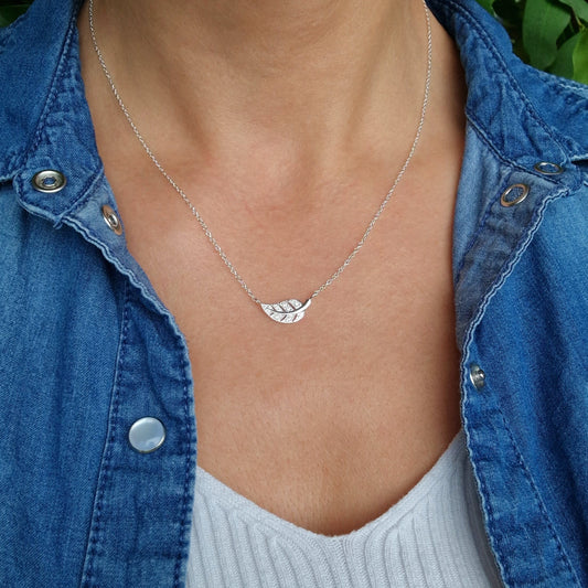 Zarte Halskette mit liegendem Blatt mit Zirkonia am Hals des Models in Nahaufnahme. Das Model trägt eine Jeansbluse und ein weißes Top, im Hintergrund ist ein grüner Busch