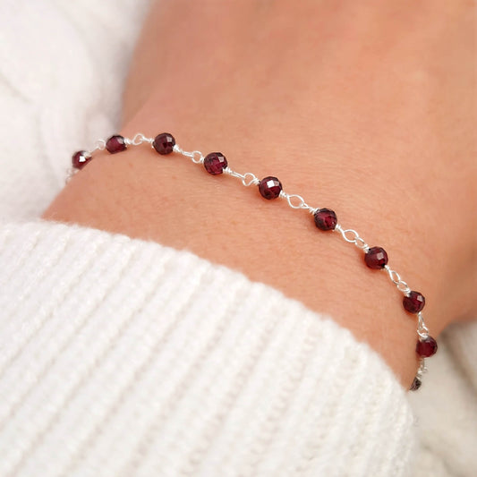 zartes Silber Armband mit bordeaux roten facettierten Rhodolit  Perlen aus Naturstein am Handgelenk des Models in seitlicher Nahaufnahme mit weißem Pulli aus Wolle