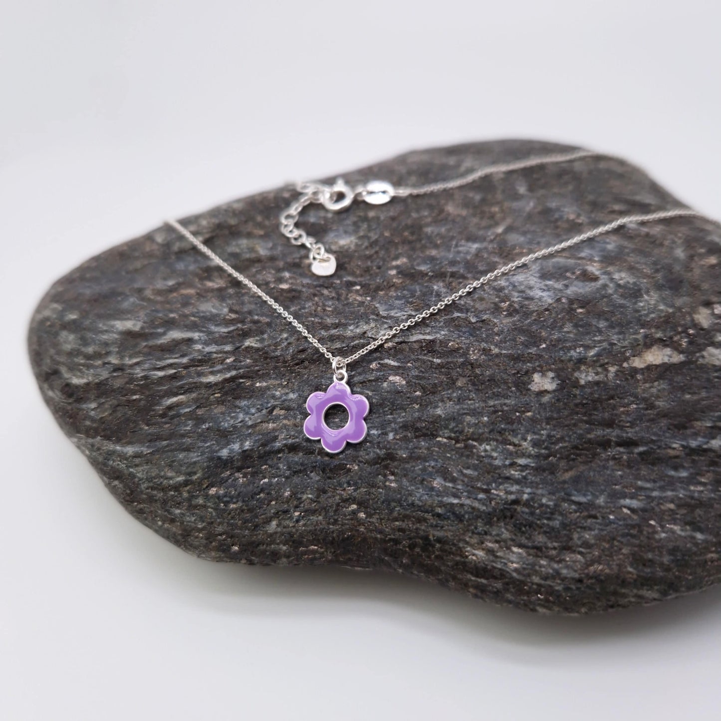 Halskette aus massivem 925 Sterling Silber mit Anhänger runde, lila emaillierte, fünfblättrige Blüte mit Loch in der Mitte und auf einem dunklen Stein drapiert und fotografiert 
Die Halskette hat ein Verlängerungskettchen mit einem kleinen Anhänger