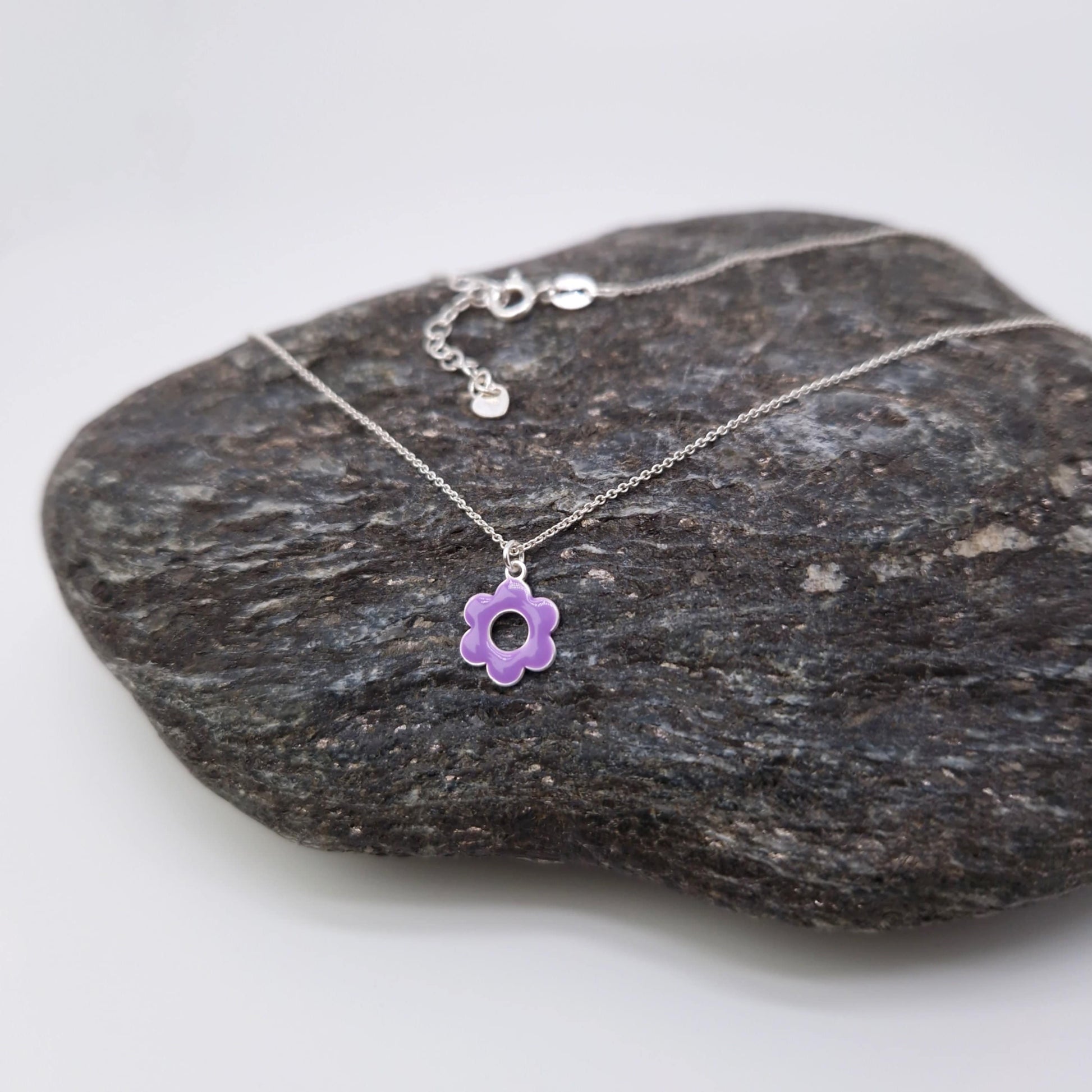 Halskette aus massivem 925 Sterling Silber mit Anhänger runde, lila emaillierte, fünfblättrige Blüte mit Loch in der Mitte und auf einem dunklen Stein drapiert und fotografiert 
Die Halskette hat ein Verlängerungskettchen mit einem kleinen Anhänger