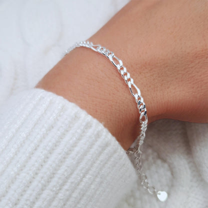 Breites Figaro Armband aus Silber am Handgelenk des Models in seitlicher Nahaufnahme, die Verlängerungskette mit kleinem Herzanhänger ist ebenfalls zu sehen. Das Model trägt einen weißen Pulli