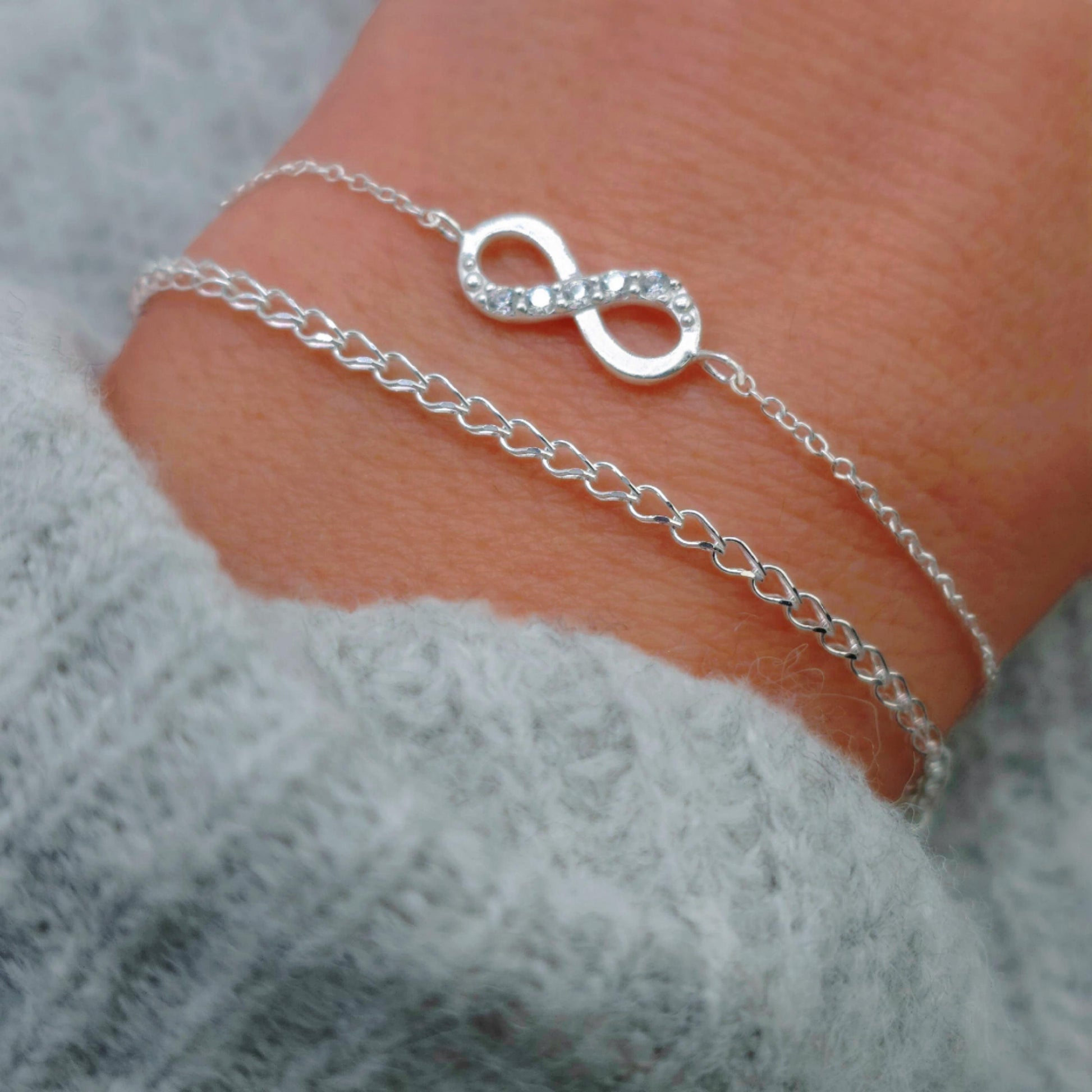 zartes  gewebtes Silber Armband am Handgelenk des Models in seitlicher Nahaufnahme mit hellgrauem Pulli kombiniert mit zartem SilbernArmband mit Unendlichkeitssymbol und Zirkoniakristallen
